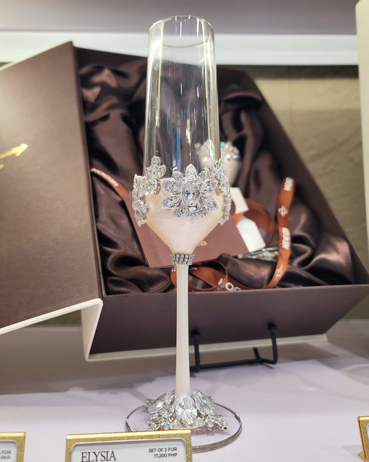 An ornate champagne flute with a white stem and silver crystal embellishments around the bowl, displayed in front of an open dark brown gift box with brown satin lining. A small sign at the bottom reads ELYSIA.