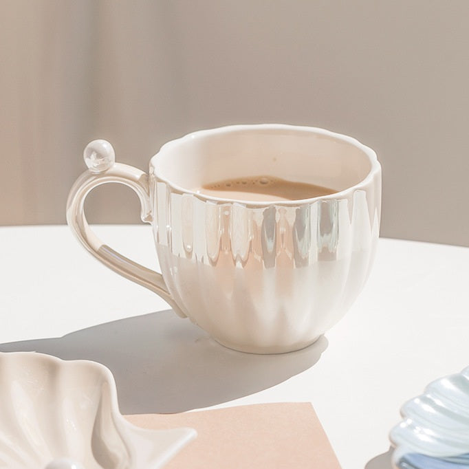 Designed to mimic the graceful curves of a seashell, each piece boasts a delicate iridescent finish that catches the light, evoking the shimmer of ocean pearls. The sculpted handle features a charming pearl-like accent,