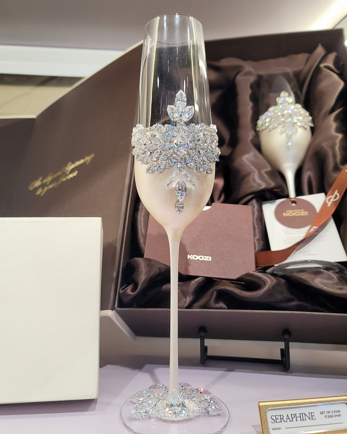 An ornate champagne flute with a white body and sparkling silver crystal embellishments, displayed in front of its brown presentation box and a second matching flute.
