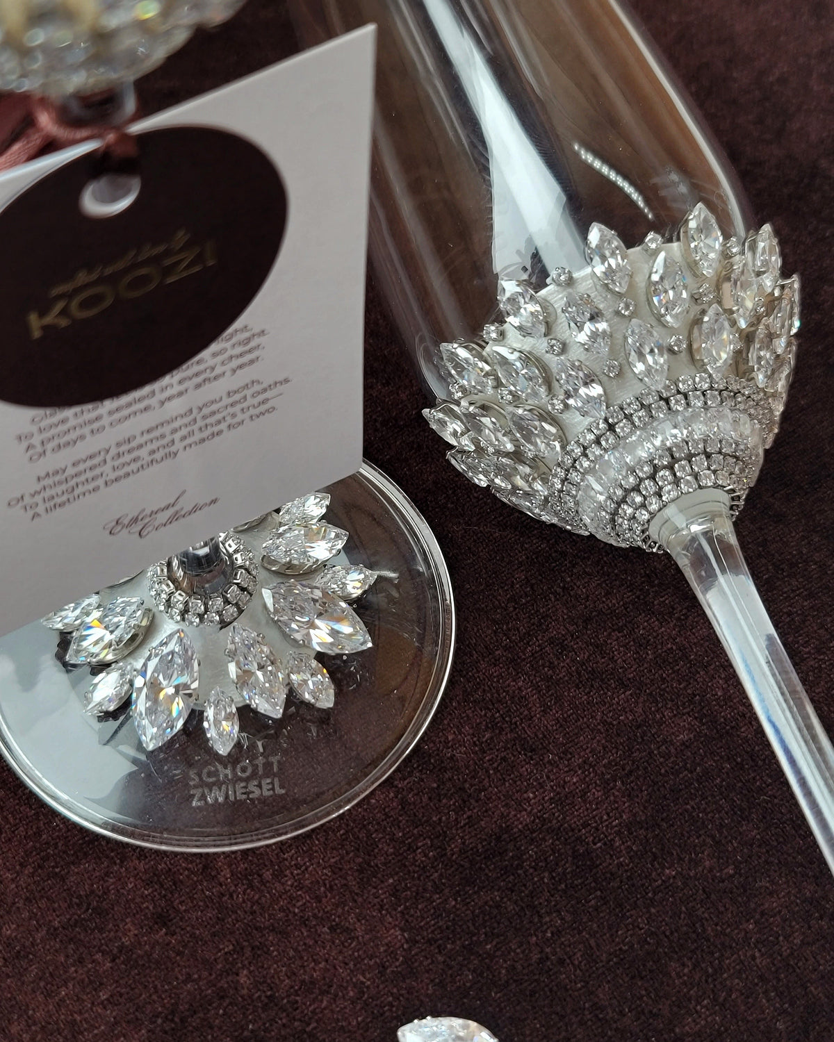 A close-up of a clear champagne flute decorated with a large silver crystal floral embellishment near the bowl, shown on a dark brown textured surface. A matching crystal-and-silver coaster sits beside it, with a branded hangtag reading KOOZI and the brand name Zwiesel visible on the coaster.