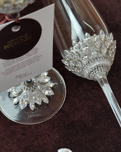 A close-up of a clear champagne flute decorated with a large silver crystal floral embellishment near the bowl, shown on a dark brown textured surface. A matching crystal-and-silver coaster sits beside it, with a branded hangtag reading KOOZI and the brand name Zwiesel visible on the coaster.