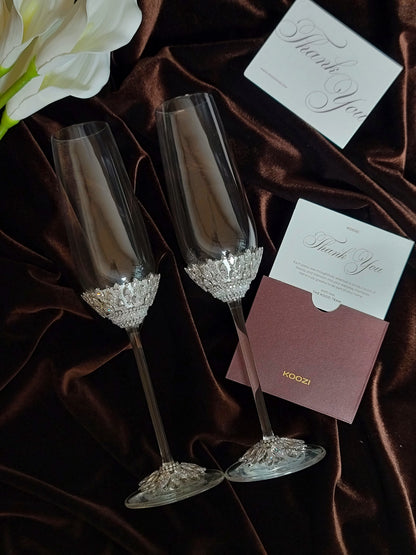 Two clear champagne flutes with ornate crystal-like embellishments near the bases, arranged on dark brown satin fabric with white floral decor and wedding thank-you cards.